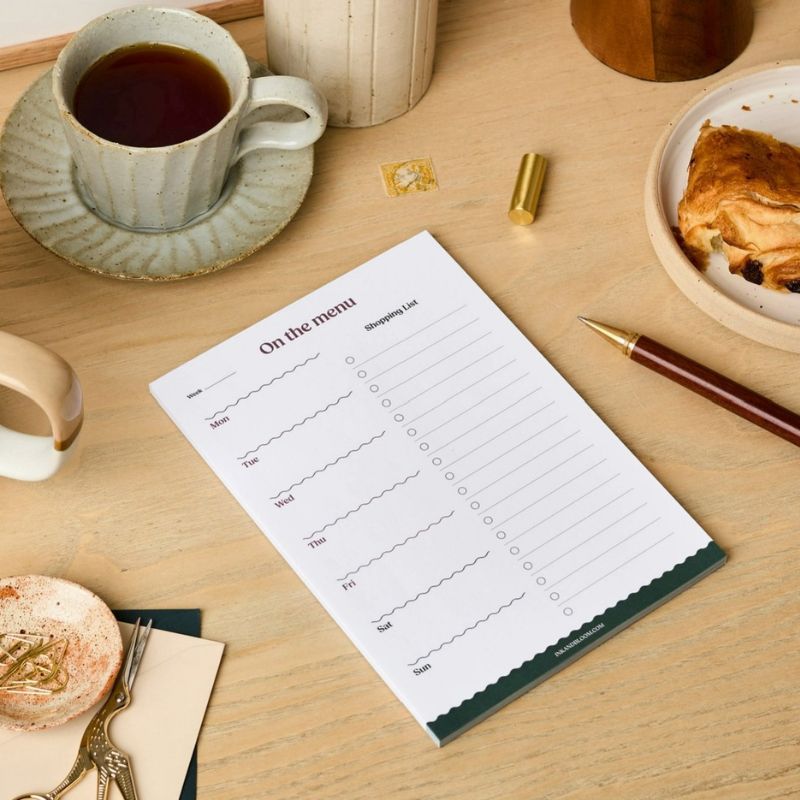 Meal Planner On the Menu Ink & Bloom A5 vierge posé en biais sur un bureau avec stylo - liste de courses et planning repas hebdomadaire
