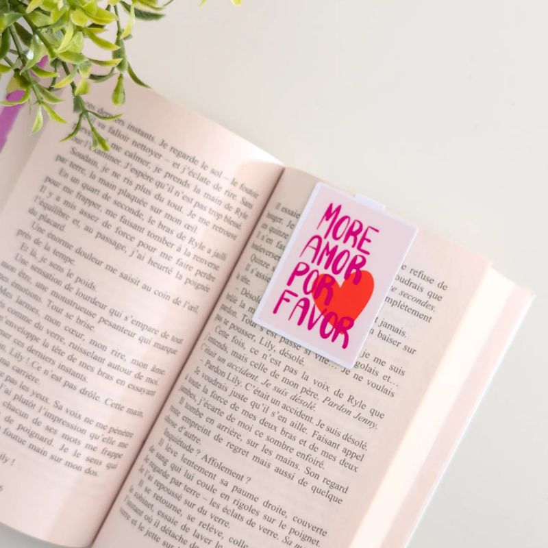 Marque-page magnétique More Amor Por Favor clipsé sur les pages d'un livre ouvert, lettering rose et cœur rouge