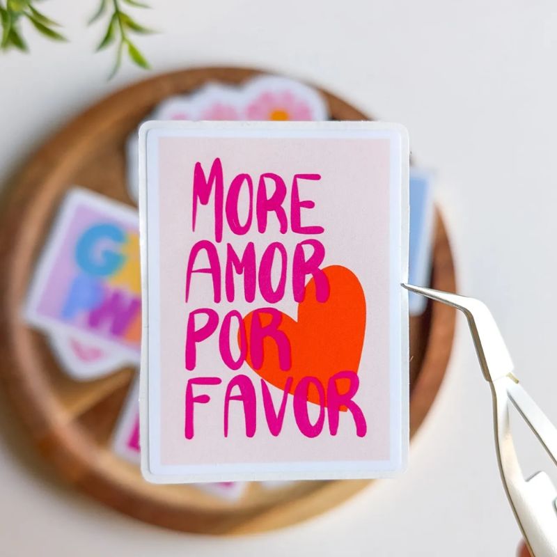 Sticker More Amor Por Favor décollé de sa planche avec une pince, lettering rose et cœur rouge