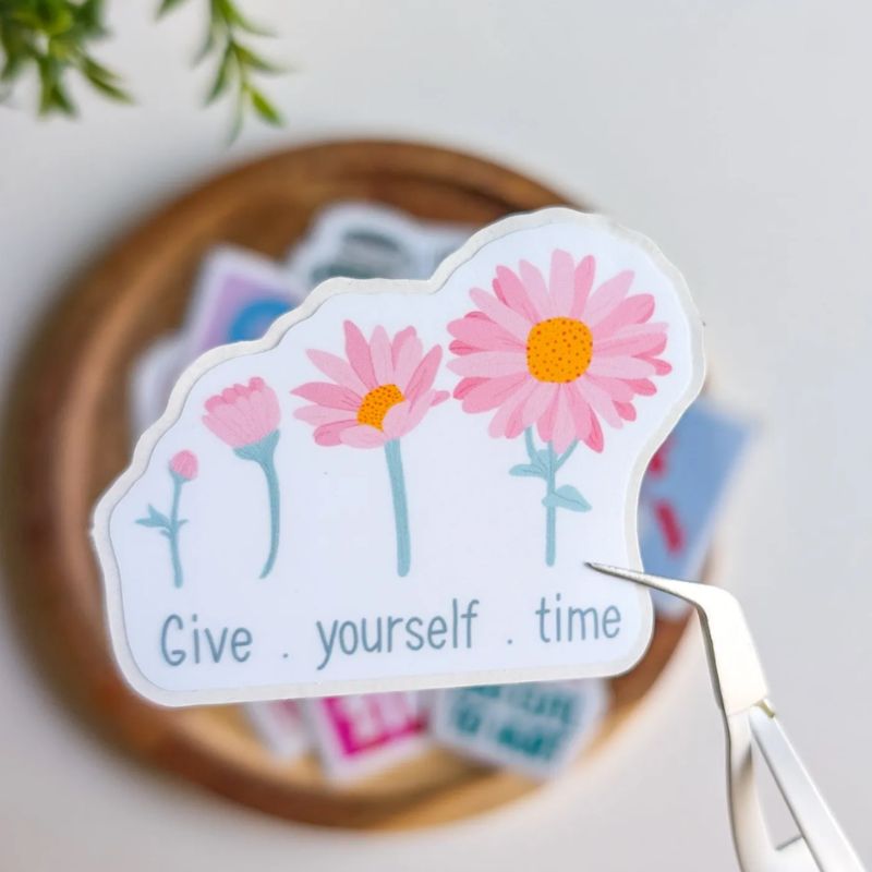 Sticker Give Yourself Time décollé de sa planche avec une pince, illustration marguerites roses