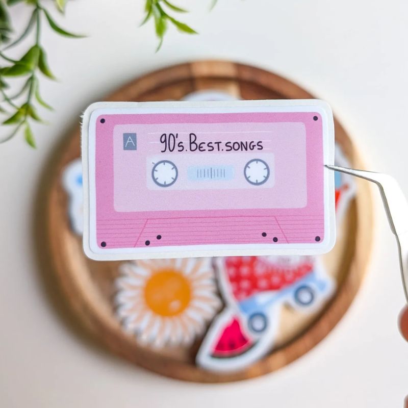 Sticker cassette rétro "90's Best Songs" décollé de sa planche avec une pince, illustration cassette audio rose