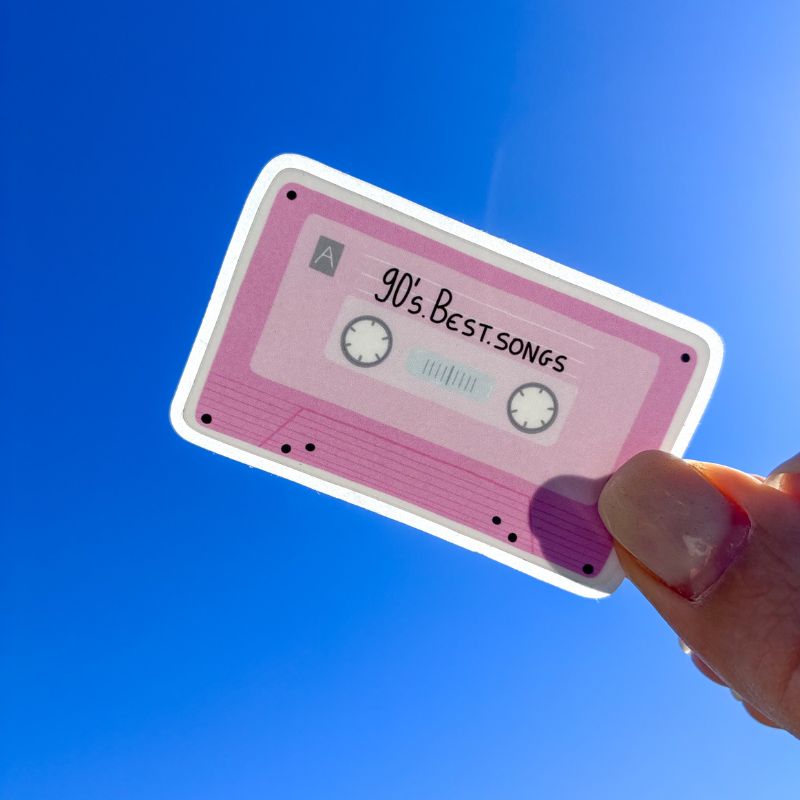 Sticker cassette rétro tenu à la main devant un ciel bleu, illustration K7 vintage rose "90's Best Songs"