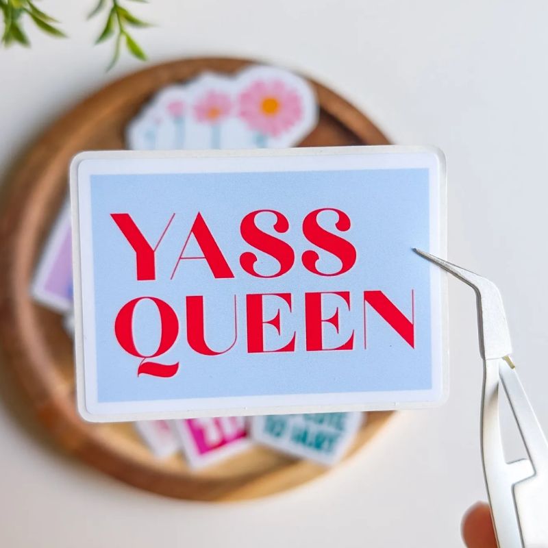 Sticker Yass Queen décollé de sa planche avec une pince, lettering rouge sur fond bleu ciel