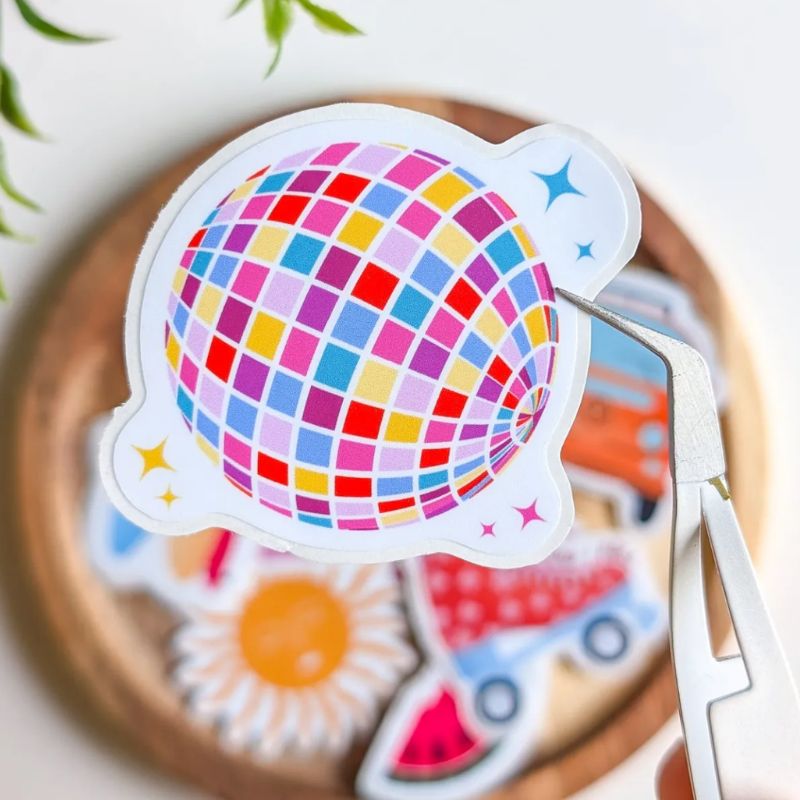 Sticker Disco Ball décollé de sa planche avec une pince, boule à facettes multicolore et étincelles