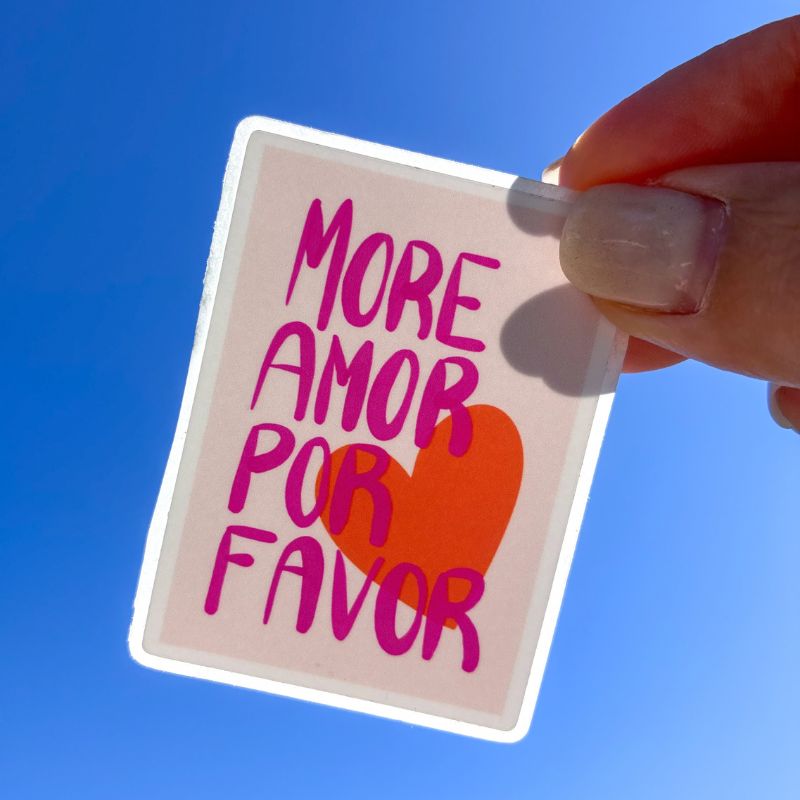 Sticker More Amor Por Favor tenu à la main devant un ciel bleu, lettering fuchsia et cœur orangé