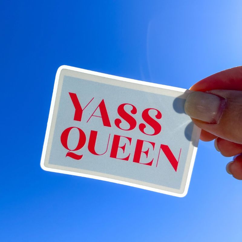 Sticker Yass Queen tenu à la main devant un ciel bleu, message de confiance en lettrage rouge
