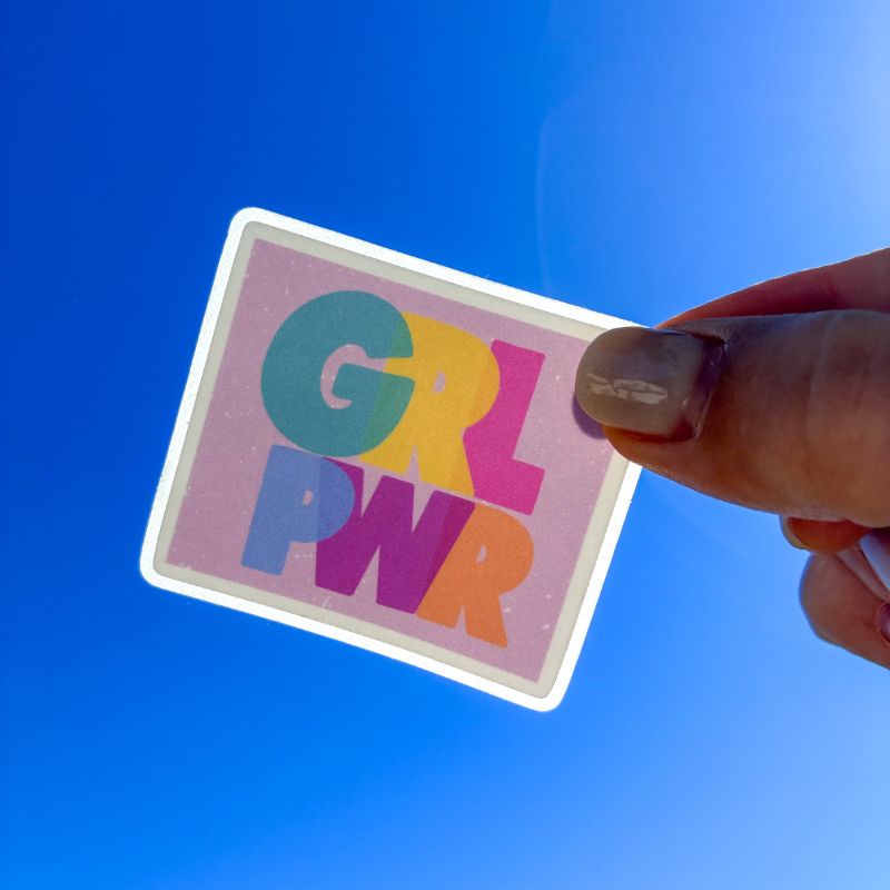 Sticker Girl Power tenu à la main devant un ciel bleu, lettering coloré féministe