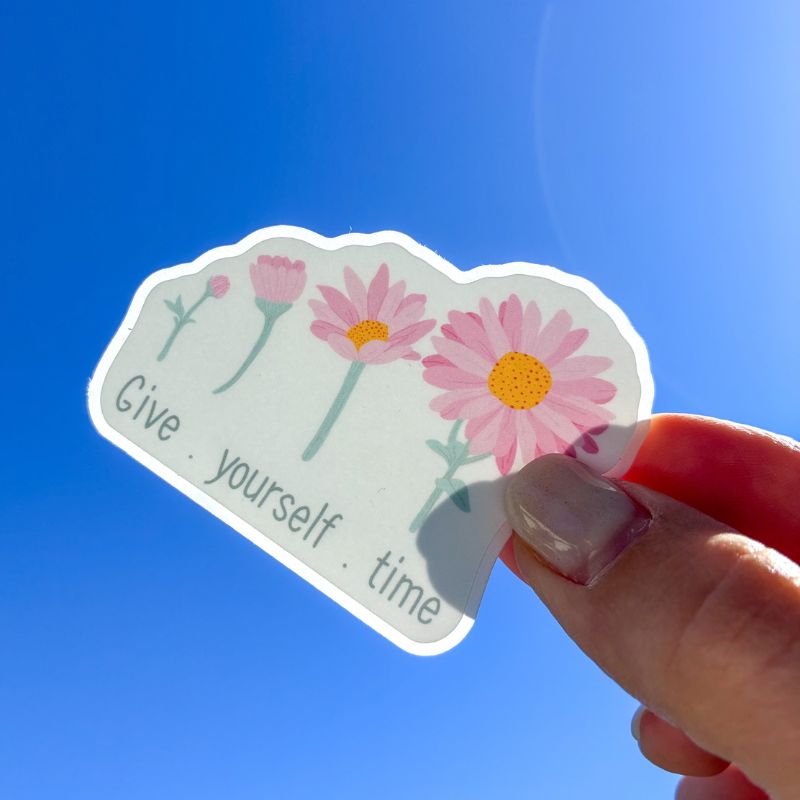 Sticker Give Yourself Time tenu à la main devant un ciel bleu, illustration marguerites roses
