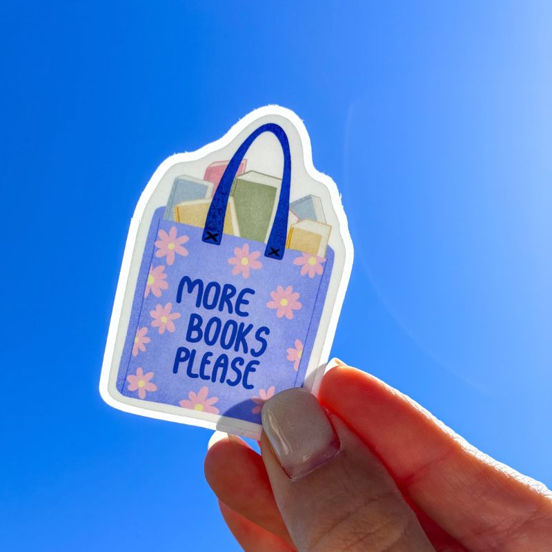 Sticker More Books Please tenu à la main devant un ciel bleu, tote bag fleuri rempli de livres